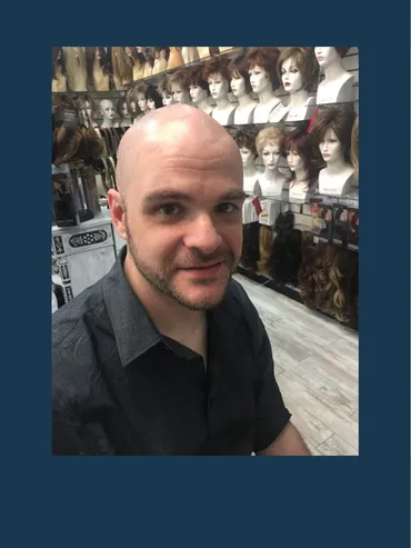 A man with bald head and black shirt in front of display of wigs.