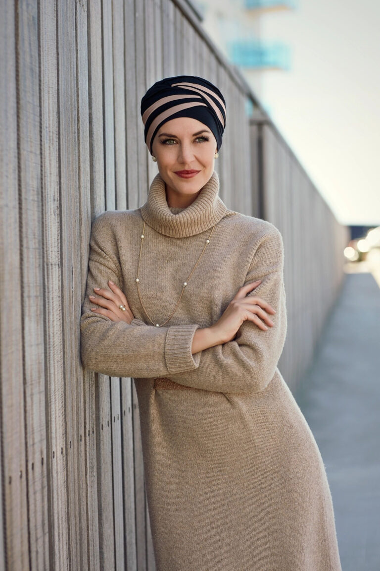 A woman in a tan coat and hat leaning on a wall.